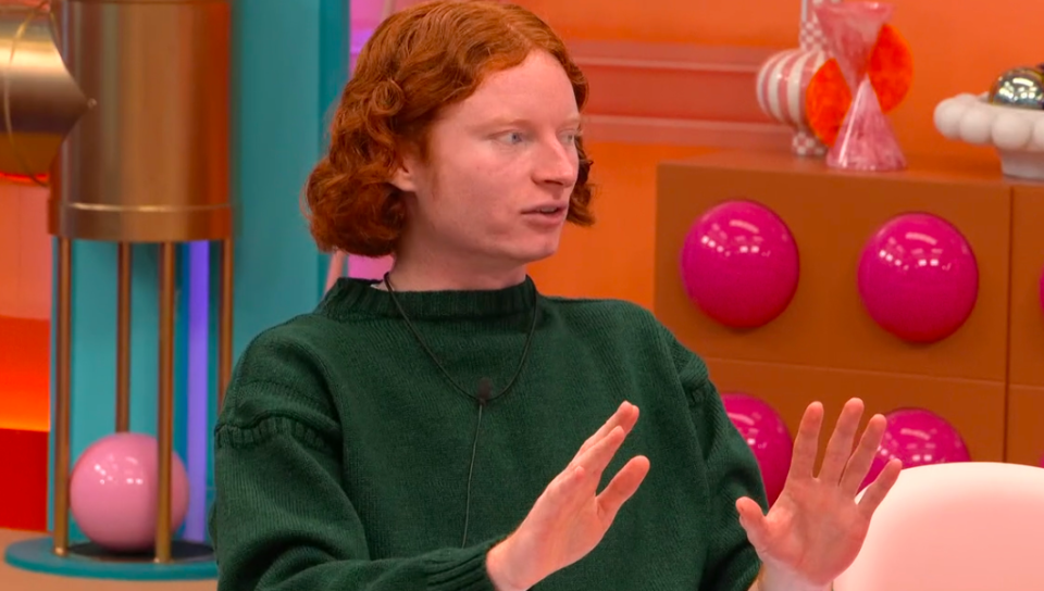 George from Big Brother in a green sweater, speaking with hands raised.