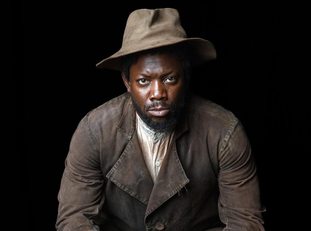 An actor, wearing a weathered brown hat and jacket, stares into the camera.