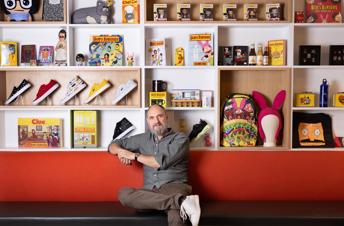 A man in a grey shirt and brown pants sits with his leg crossed on a red bench. Bob's Burgers memorabilia lines the walls.