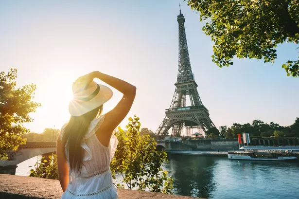 Young woman tourist in sun hat and white dress standing in front of Eiffel Tower in Paris at sunset.