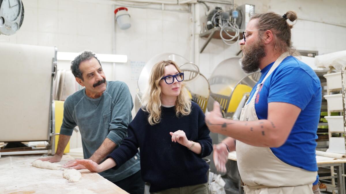 Two men flank a woman standing at a table with dough on the surface.