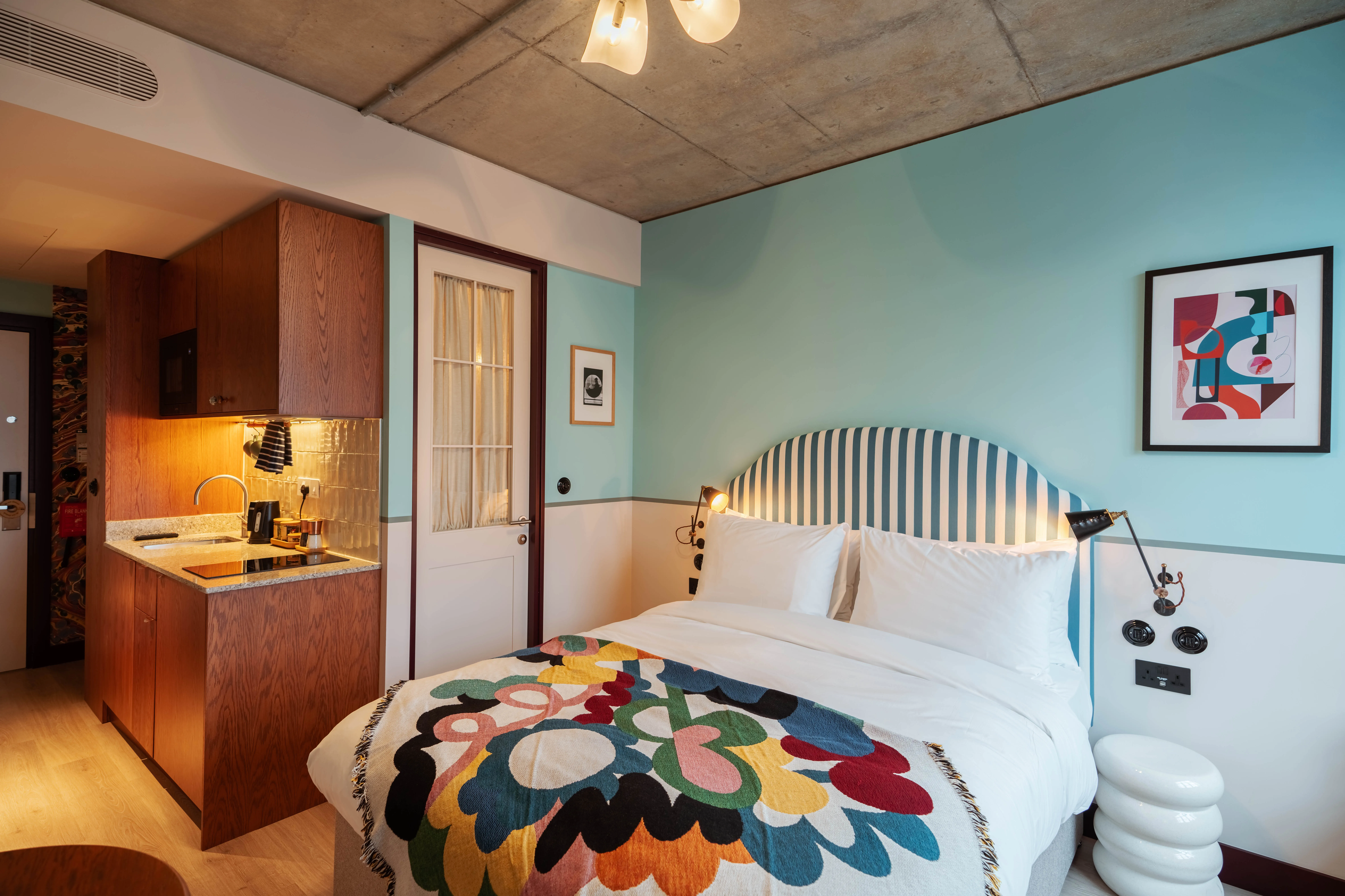 Hotel bedroom with a bed, kitchenette, and artwork.