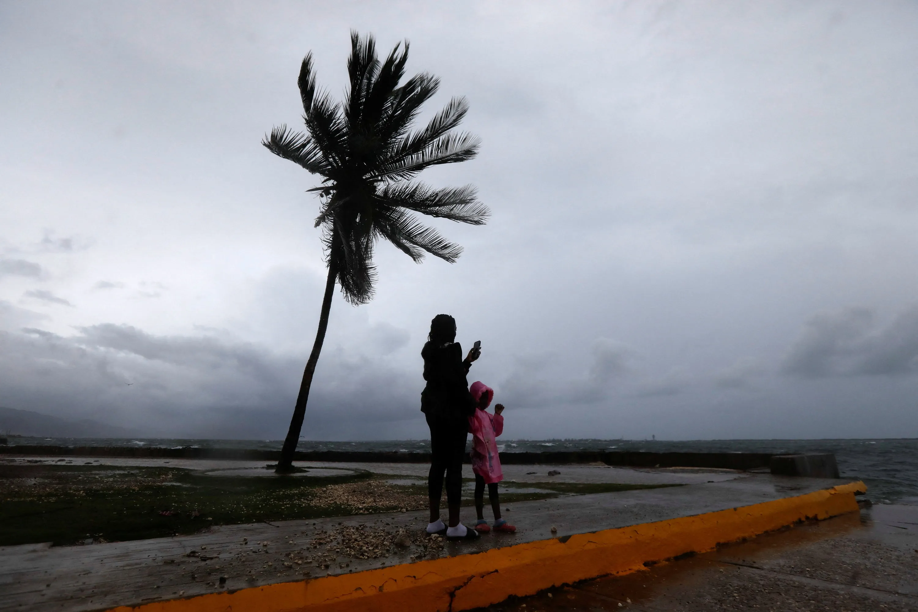 A woman and child stand on the Kingston waterfront as Hurricane Melissa approaches, with a palm tree swaying in the wind and turbulent water in the background.