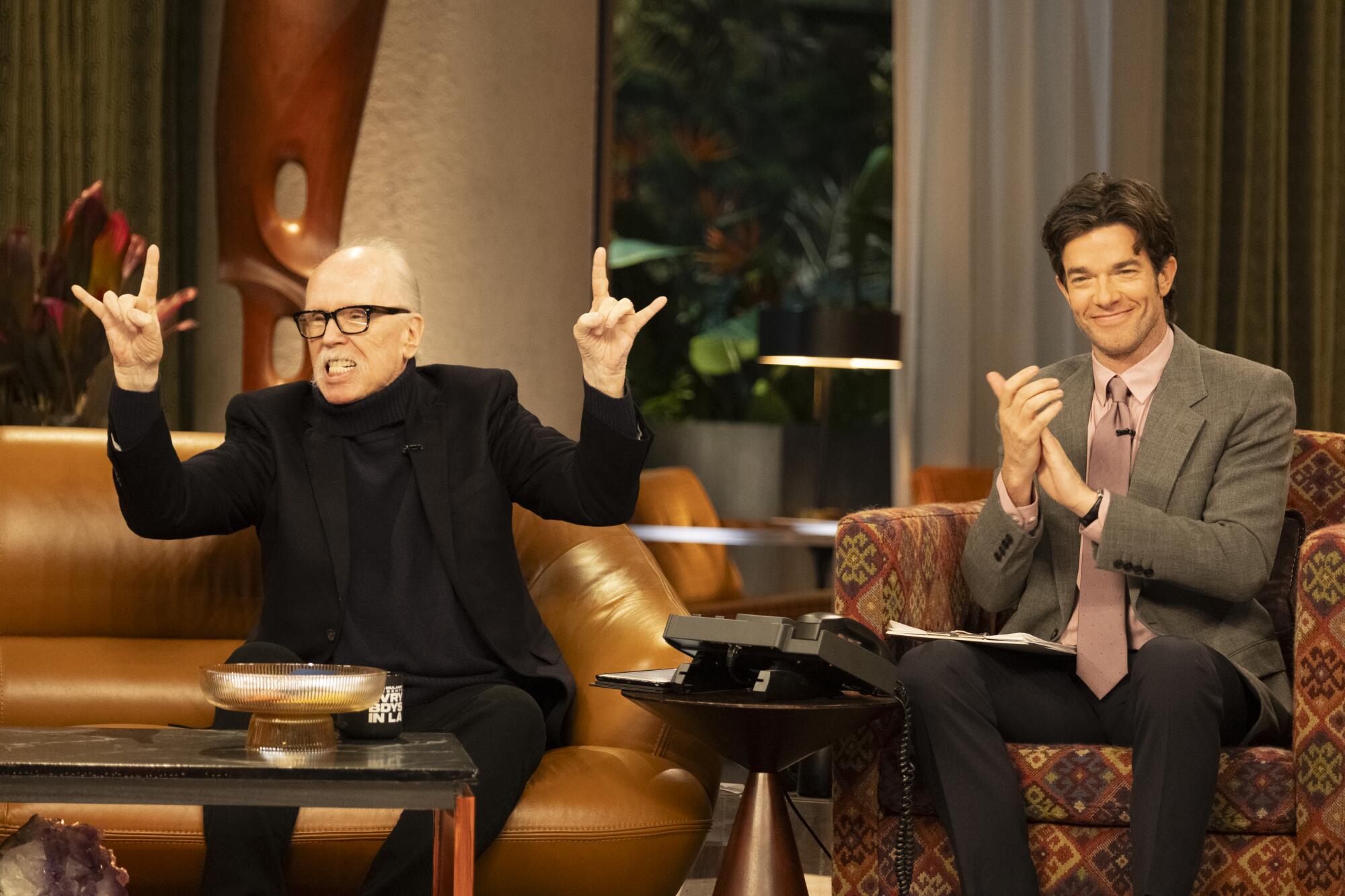 A man in black appears as a guest on a streaming series with a smiling host.