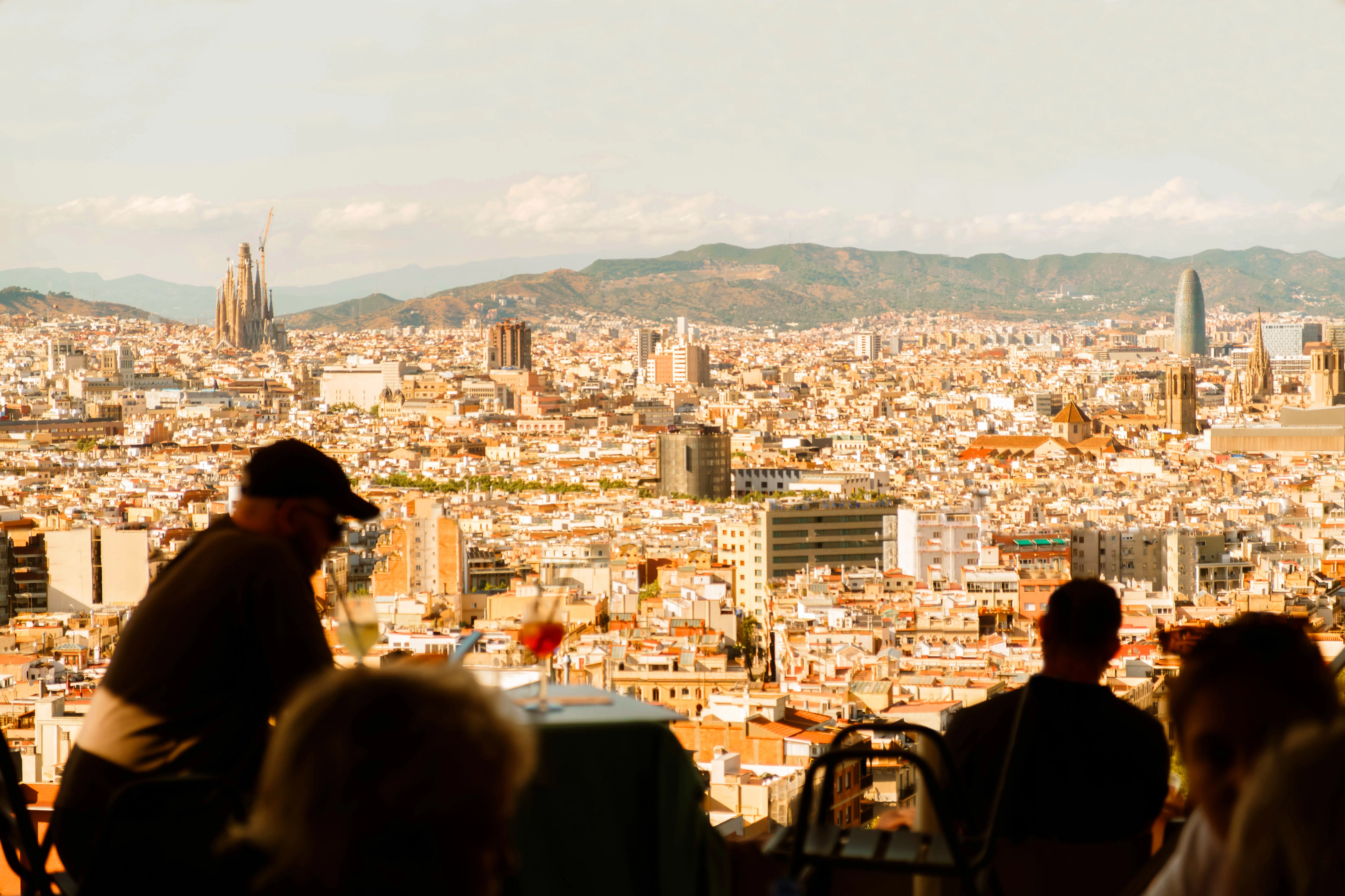 People at a rooftop bar overlooking the Barcelona skyline with Sagrada Familia and Torre Glòries visible in the background at sunset.