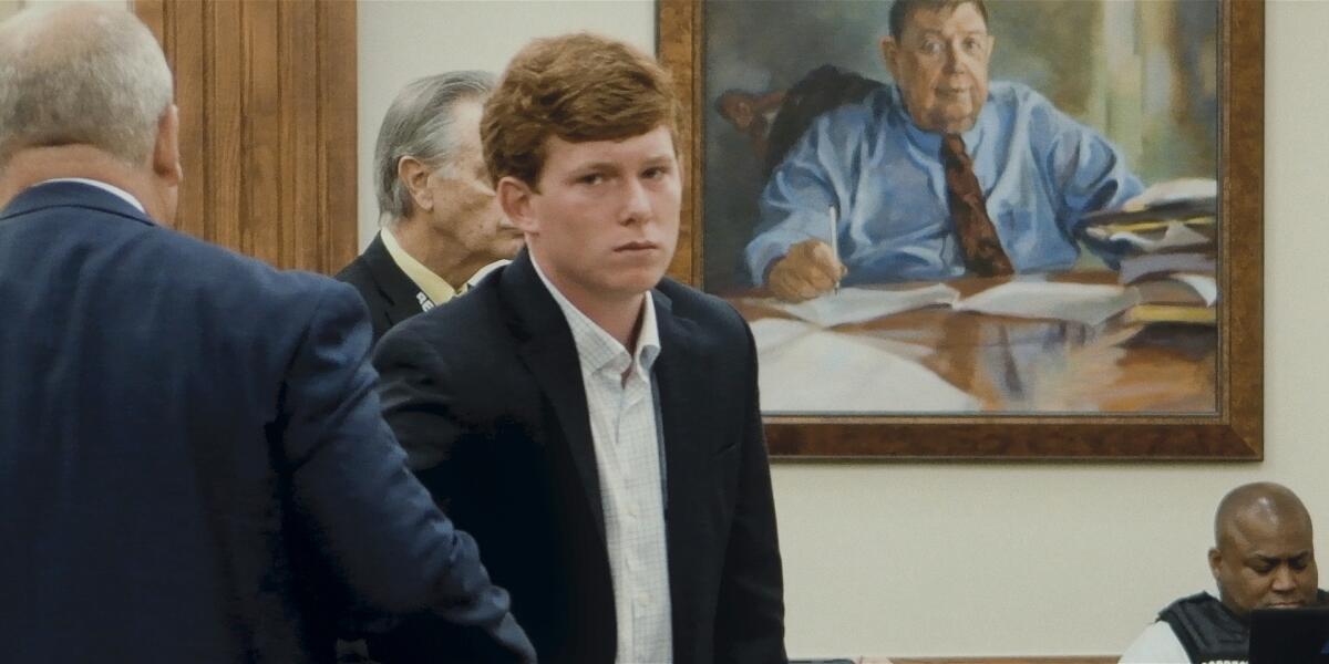 A photo of a young man in a suit standing in a courtroom.