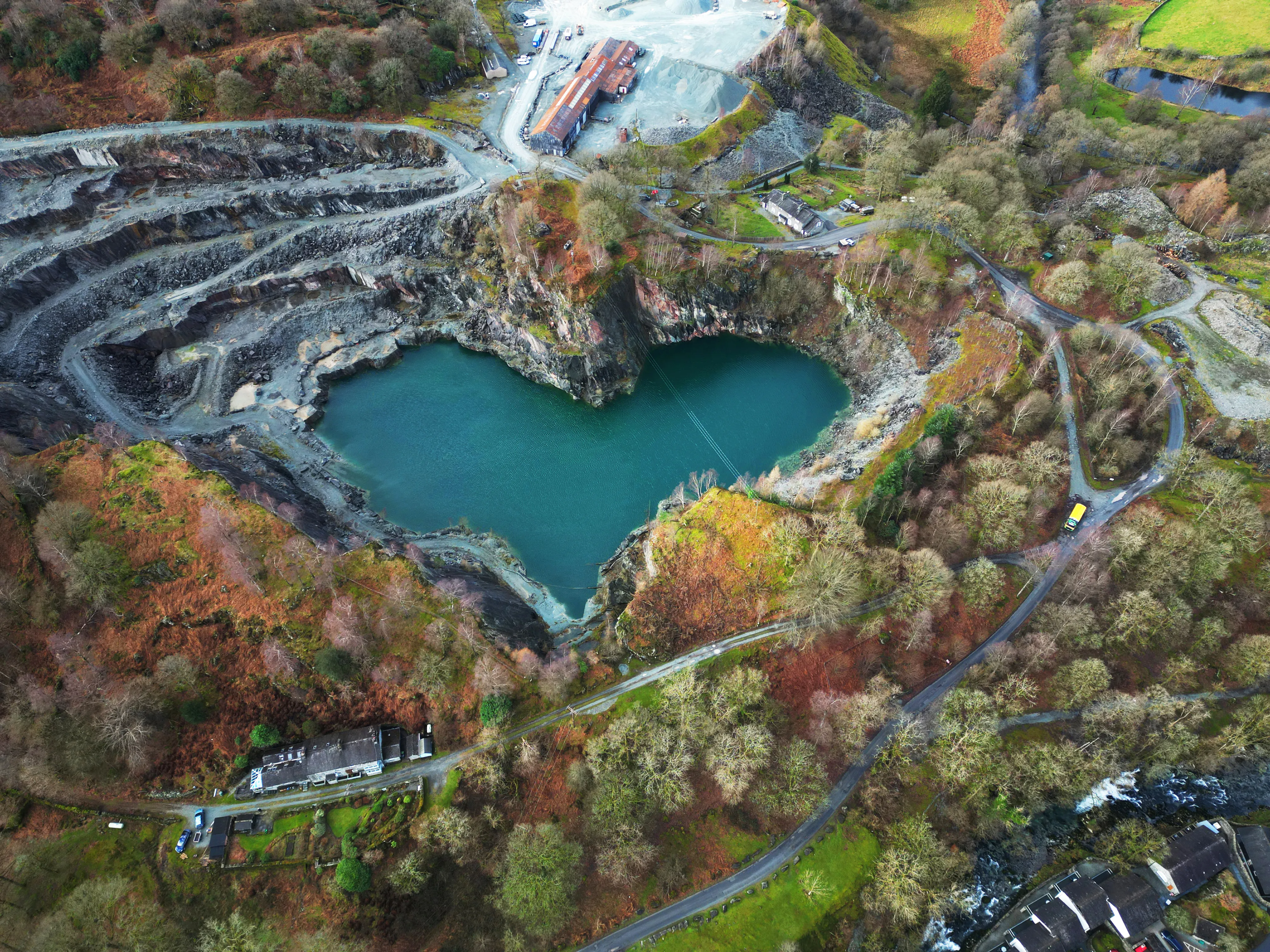 Aerial view of the heart-shaped turquoise lake at Elterwater Quarry, surrounded by trees and quarry buildings.