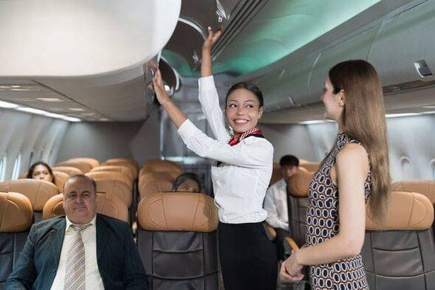 Woman flight attendant and female tourist packing travel bag in overhead baggage locker in airplance cabin