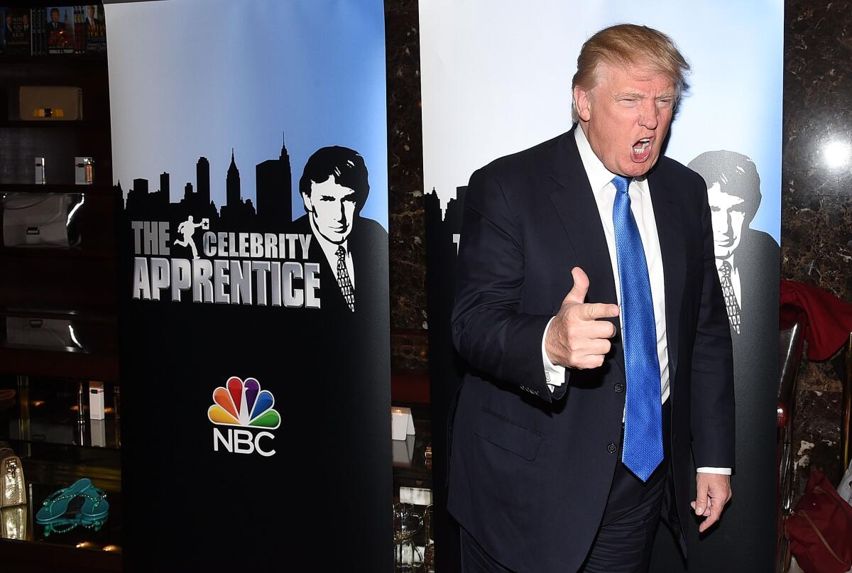 A man in a dark suit and blue tie stands with his mouth open in front of a poster for "The Celebrity Apprentice."