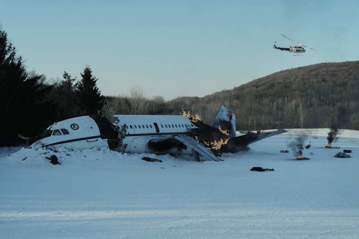 A plane on a snowy field, in flames and broken apart. A helicopter flies overhead.