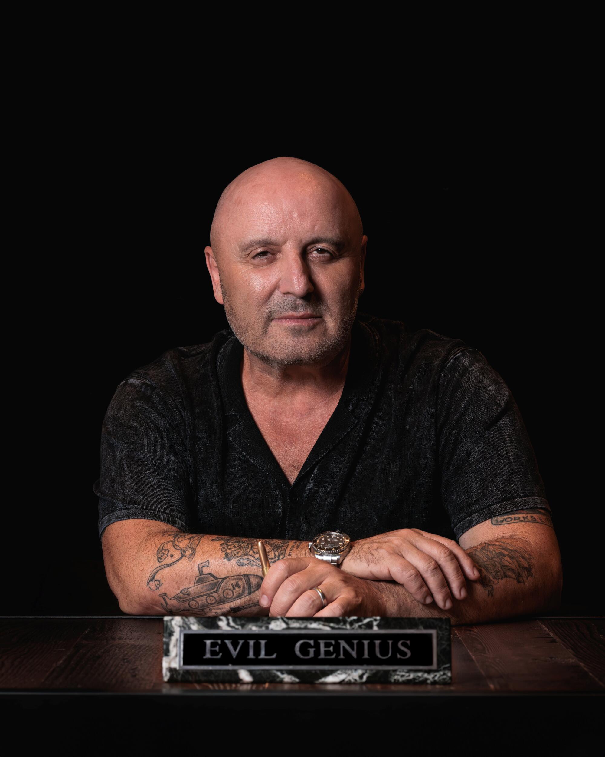 A bald man in a black sitting at a desk with a placard that says "evil genius."