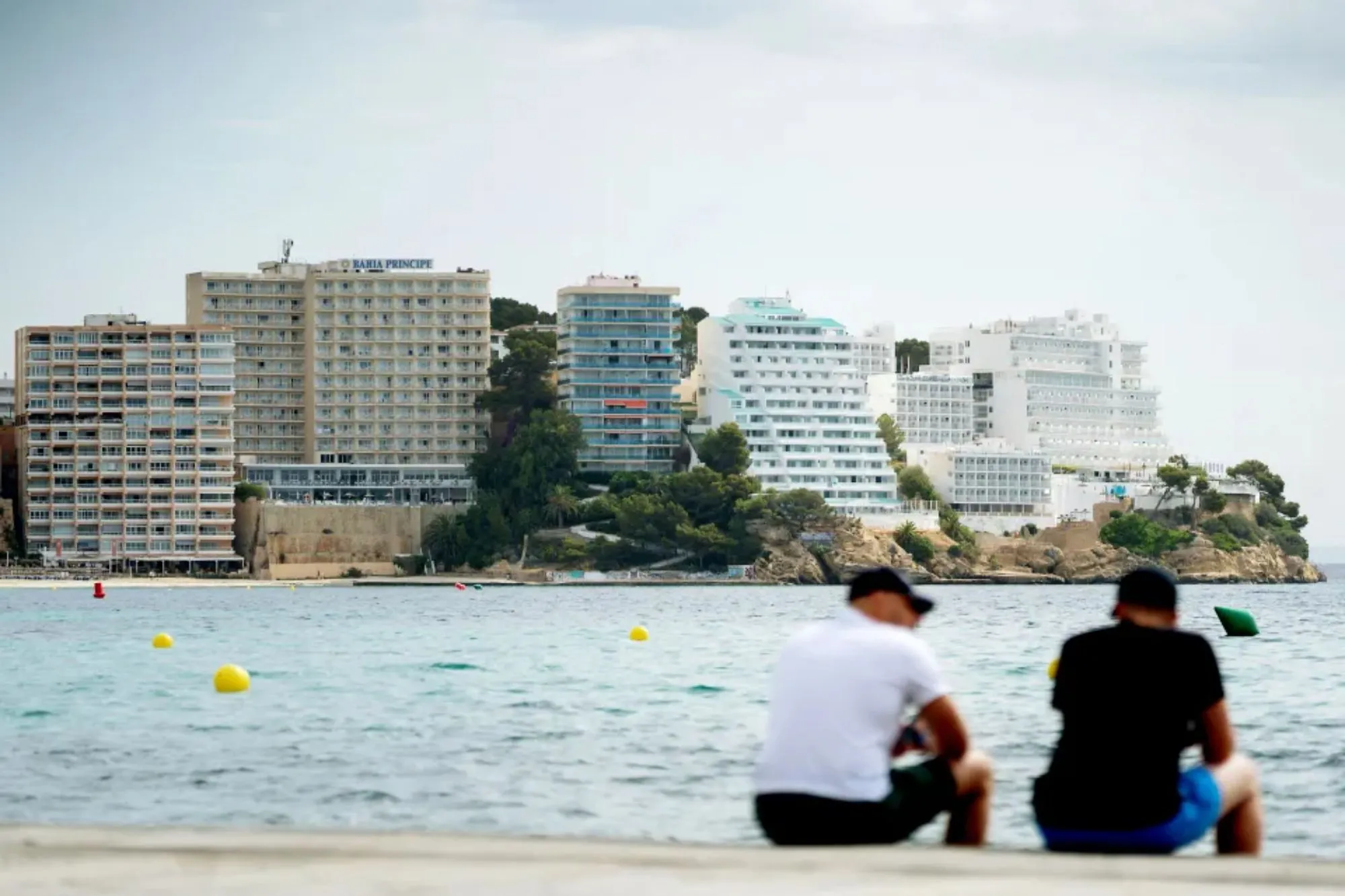 An image collage containing 1 images, Image 1 shows Two men sitting by the water overlooking a coastline of hotels