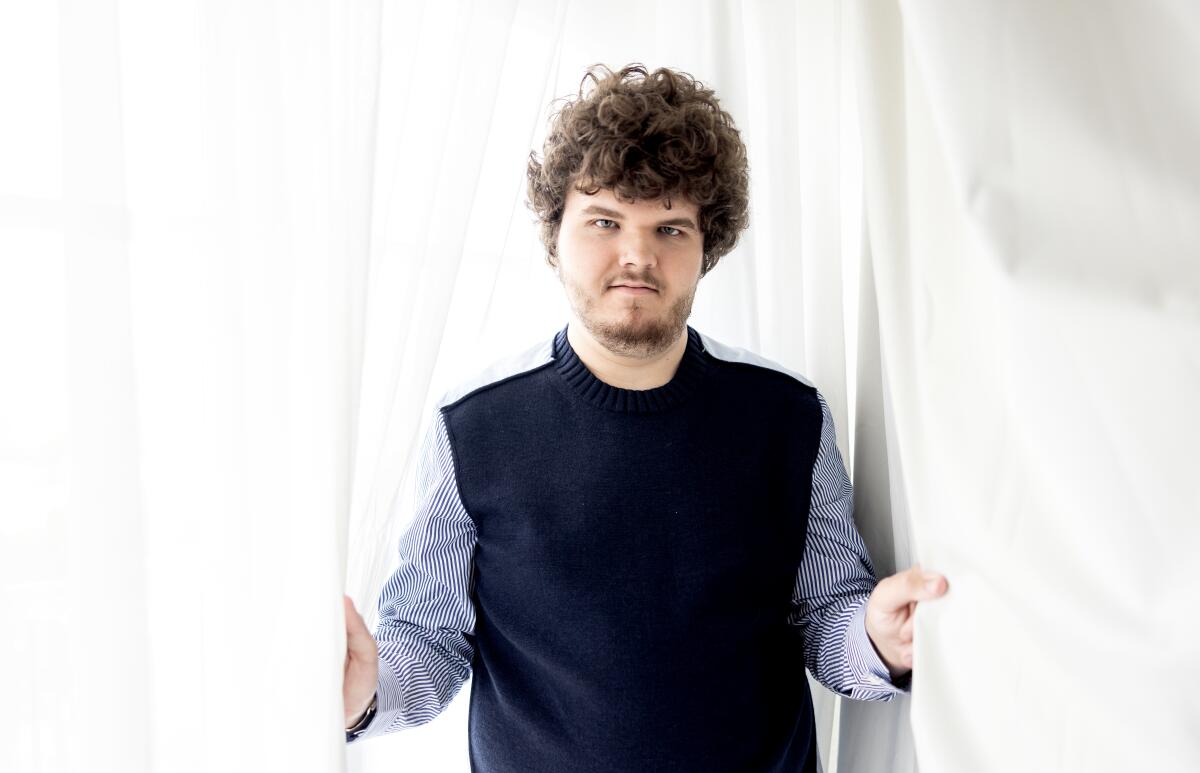 A young man with short, curly hair holds onto curtain panels while posing for a photo
