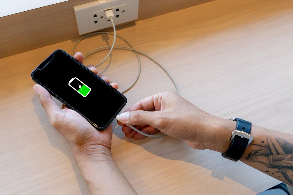 Person's hands holding a smartphone while plugging in a charger, with a green battery icon visible on the phone screen.