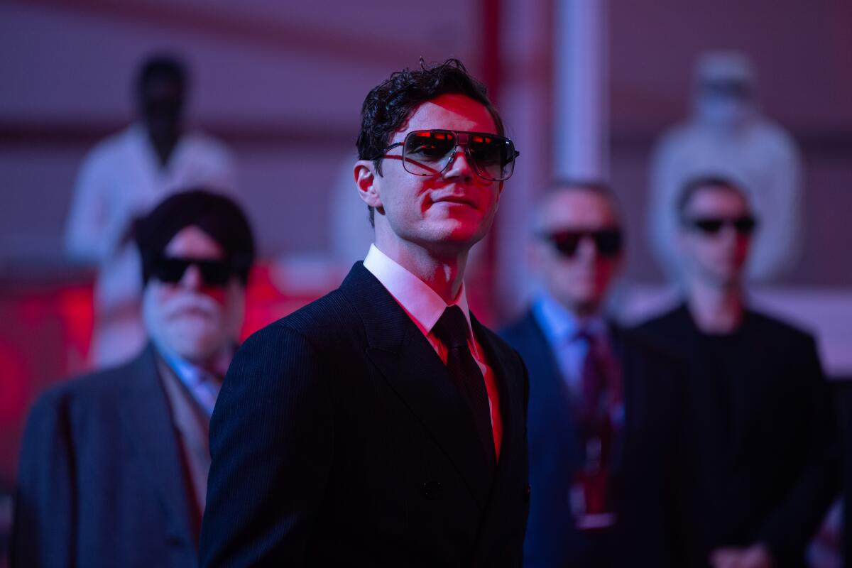 Evan Peters wears a suit and sunglasses. Behind him are other men in suits and sunglasses.