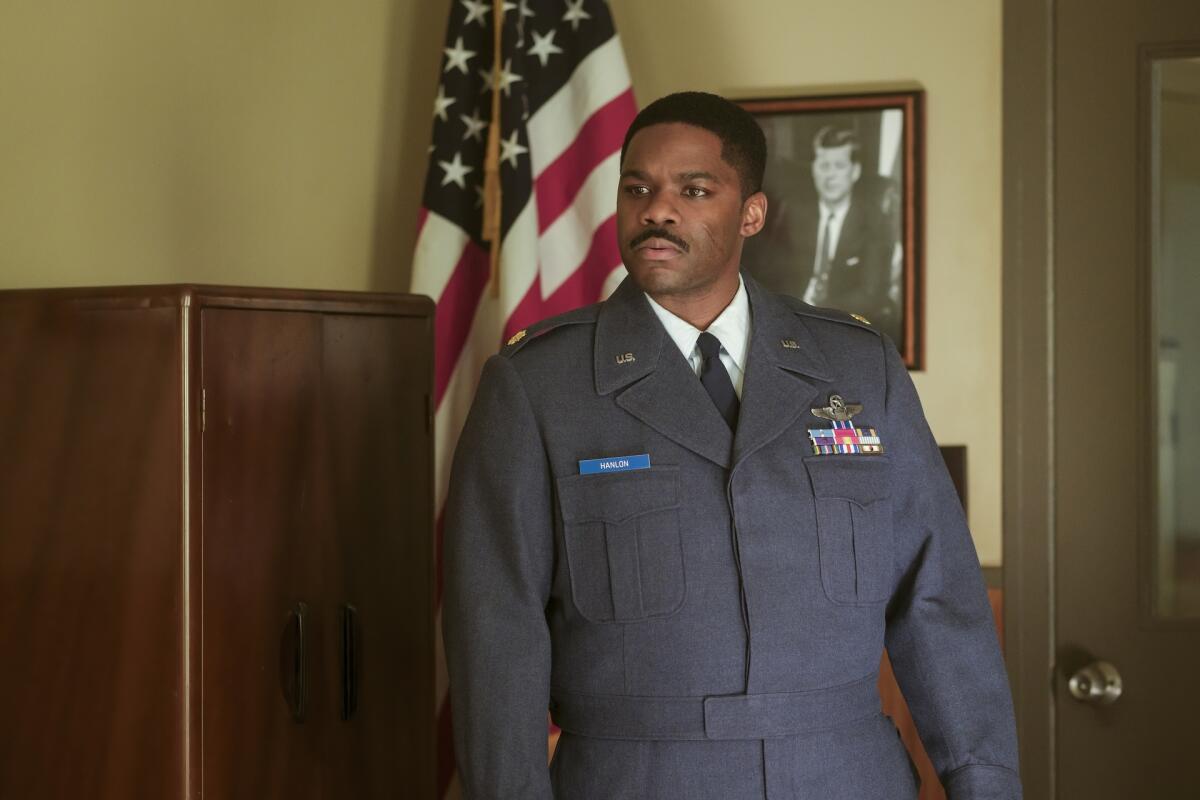 A man in a blue grey military uniform stands near a cabinet with an American flag and a photo of JFK behind him.