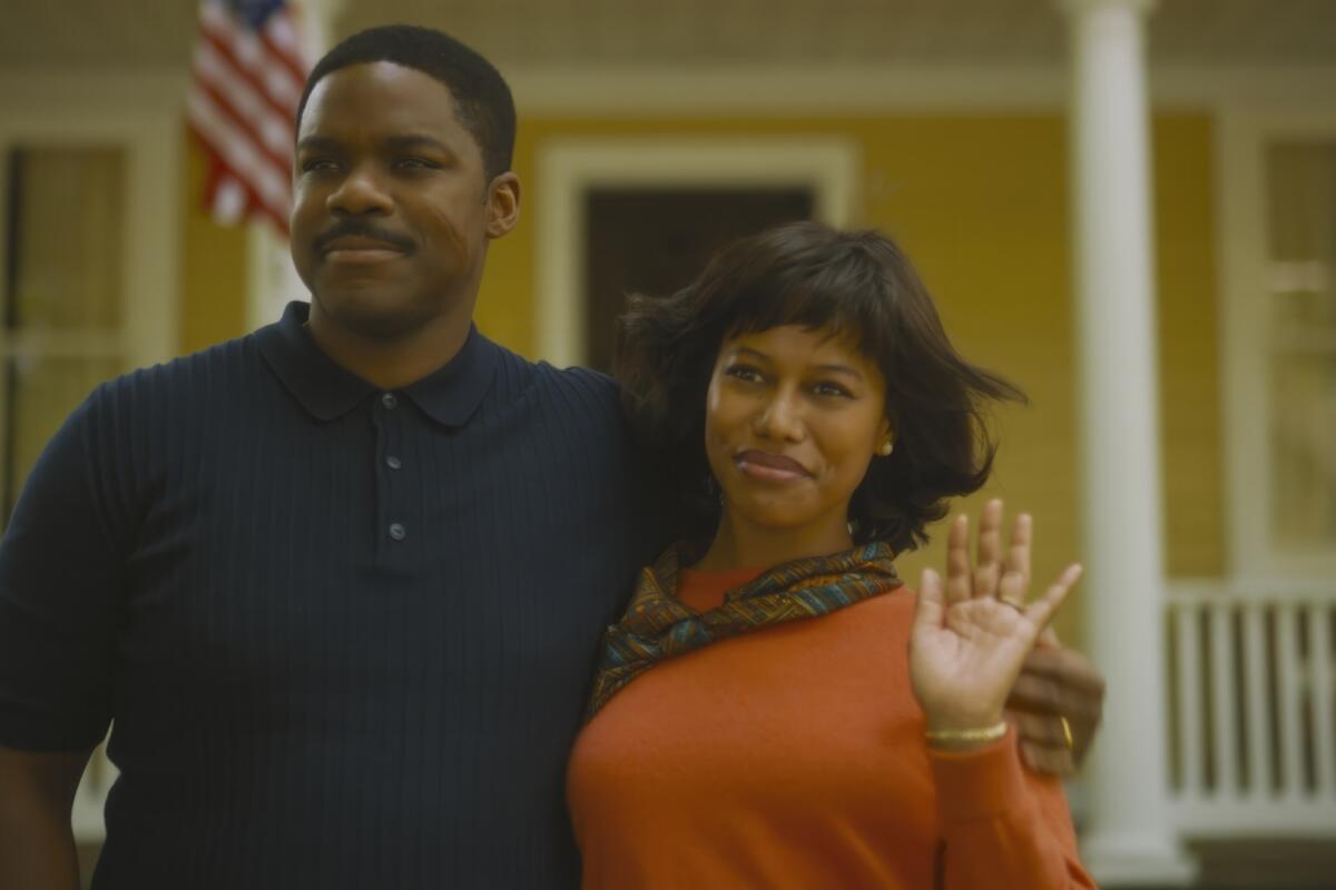 A man embracing a woman by the shoulders who waves with her hand as they stand in front of a yellow house.