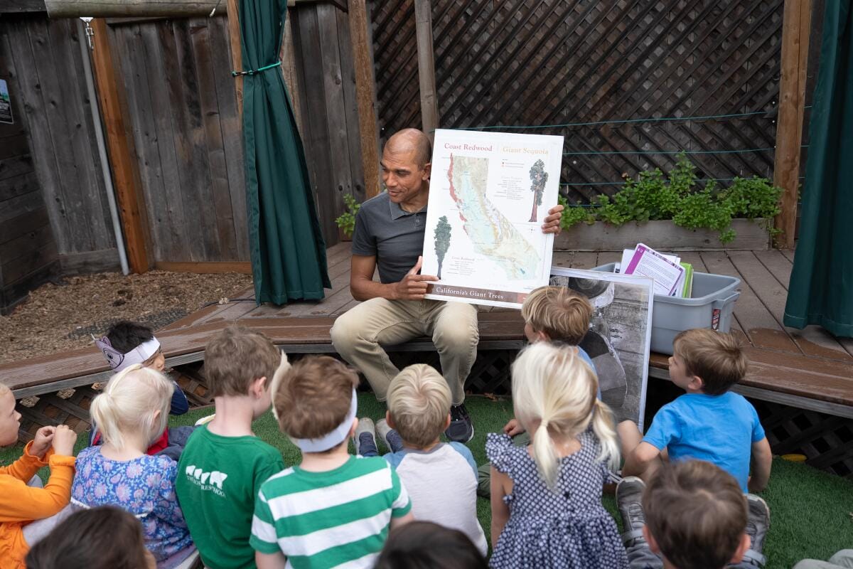 A park ranger holding an educational sign sits before a small group.
