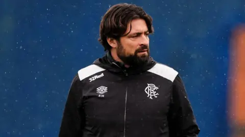 PA Media Russell Martin looks off to his left. He has black hair, combed in a middle parting, and a beard. He is wearing a black jacket with the Rangers crest and Umbro logo on it. He has been photographed in the rain against a blue backdrop.
