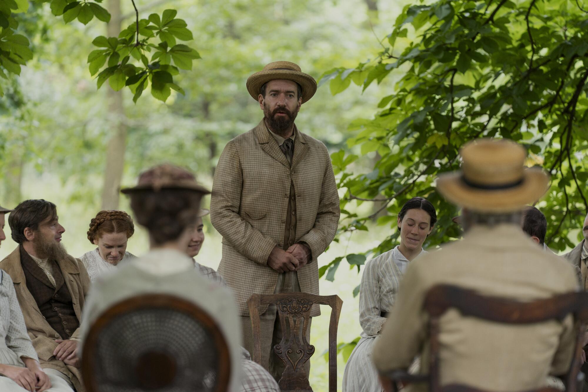 A man in a straw hat and dirty jacket stands in front of a chair surrounded by people.