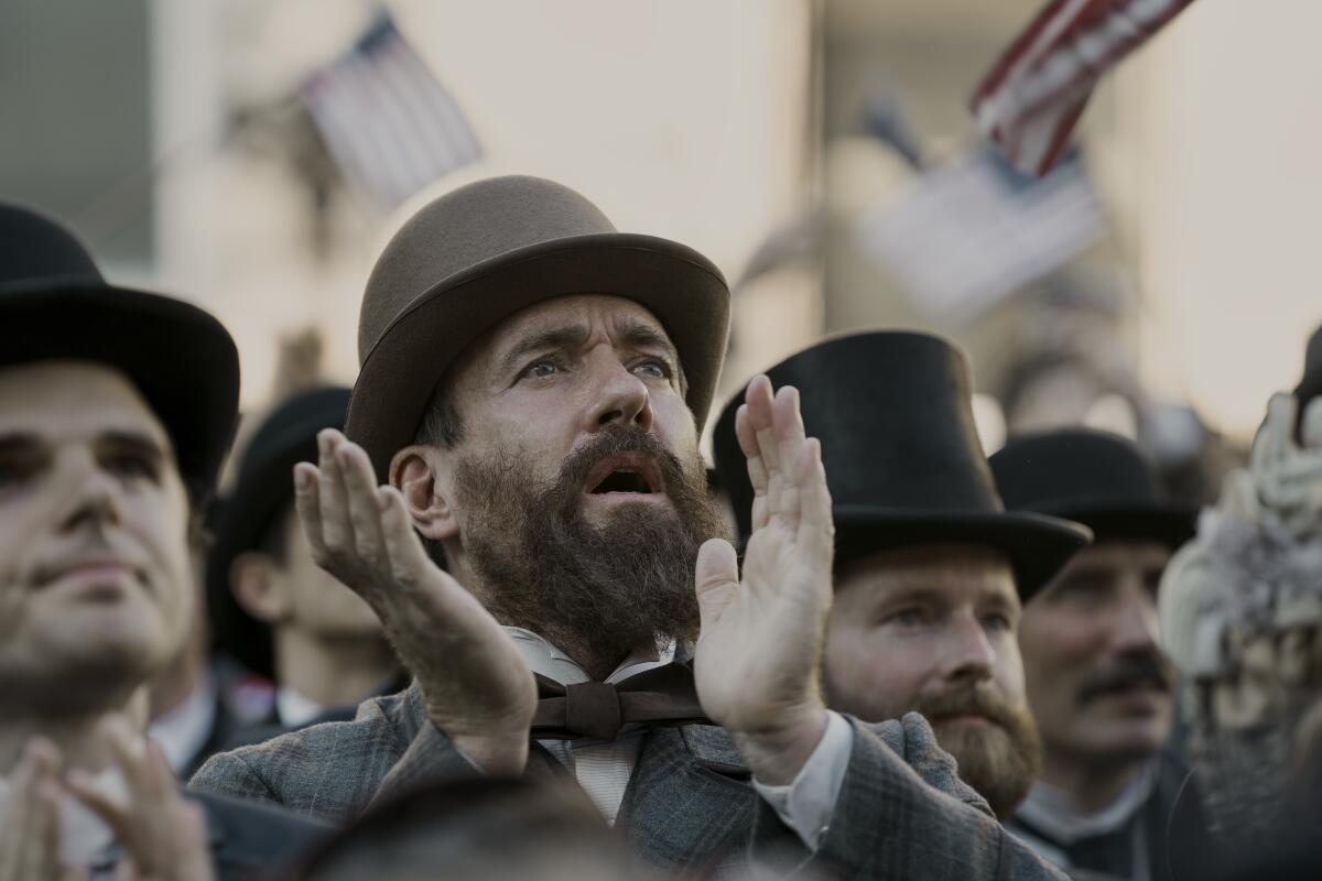 A bearded man in a tan bowler hat standing in a crowd mid-applause.