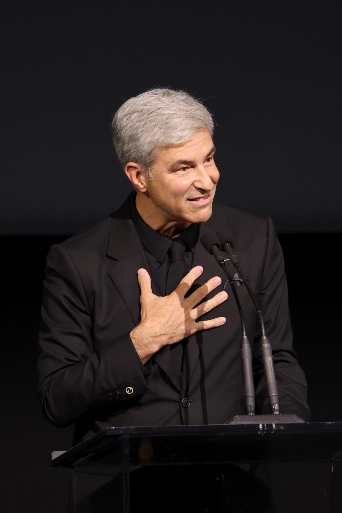 A man in a black suit speaks at microphones