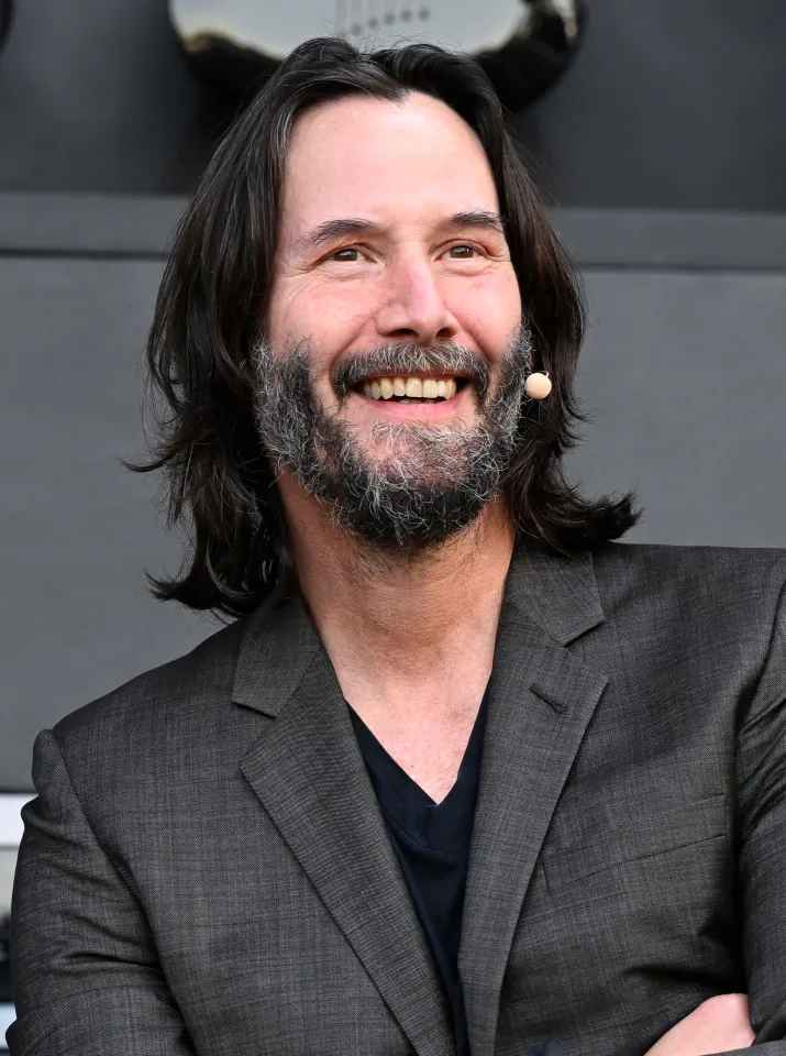 Keanu Reeves smiling, wearing a dark suit jacket and black t-shirt, with long dark hair and a gray beard, attending the Culinary Stage at BottleRock Napa Valley Music Festival.
