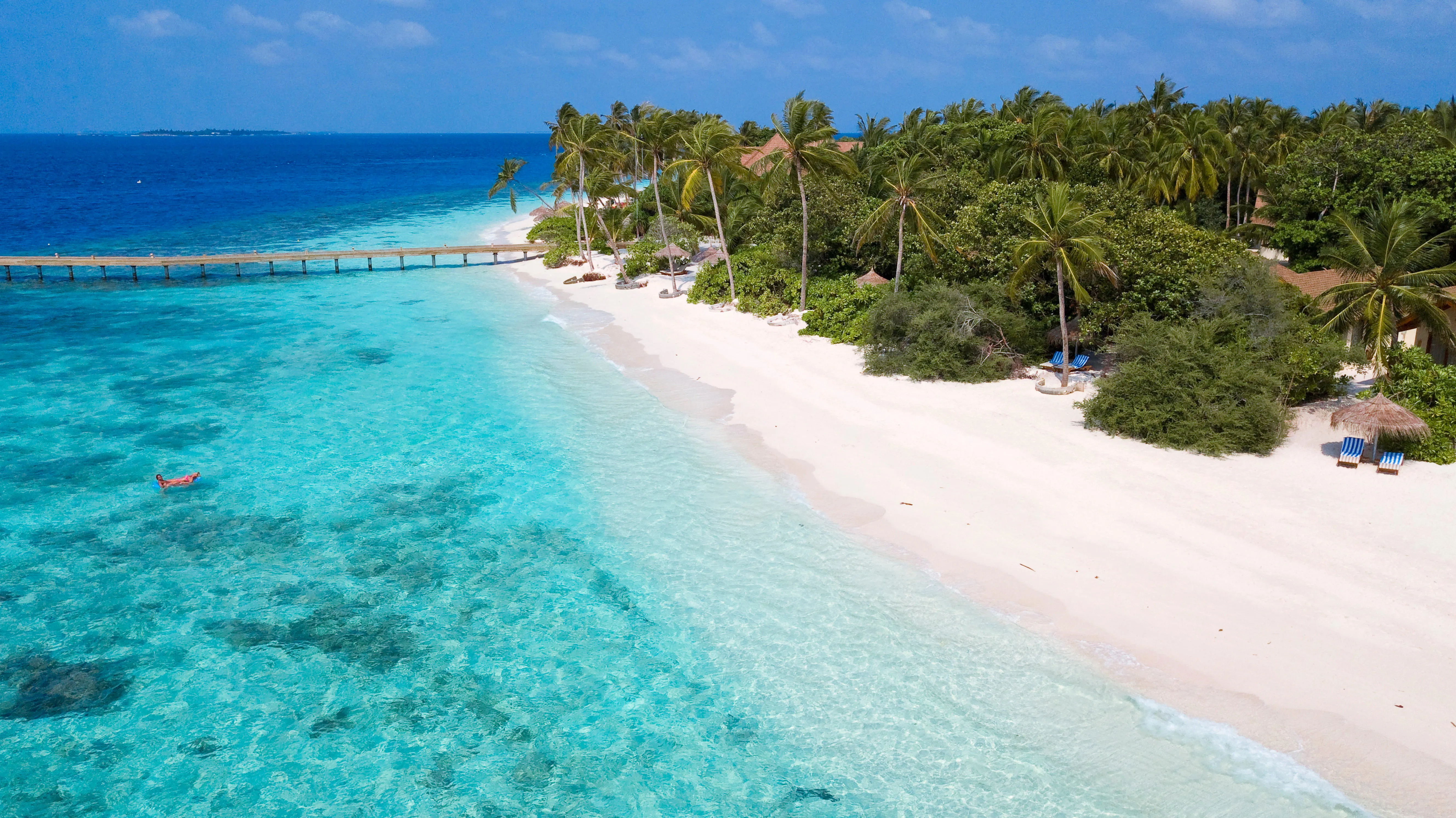 Aerial view of Filaidhoo island in Maldives, with white sandy beaches, palm trees, clear turquoise water, and a wooden pier extending into the ocean.